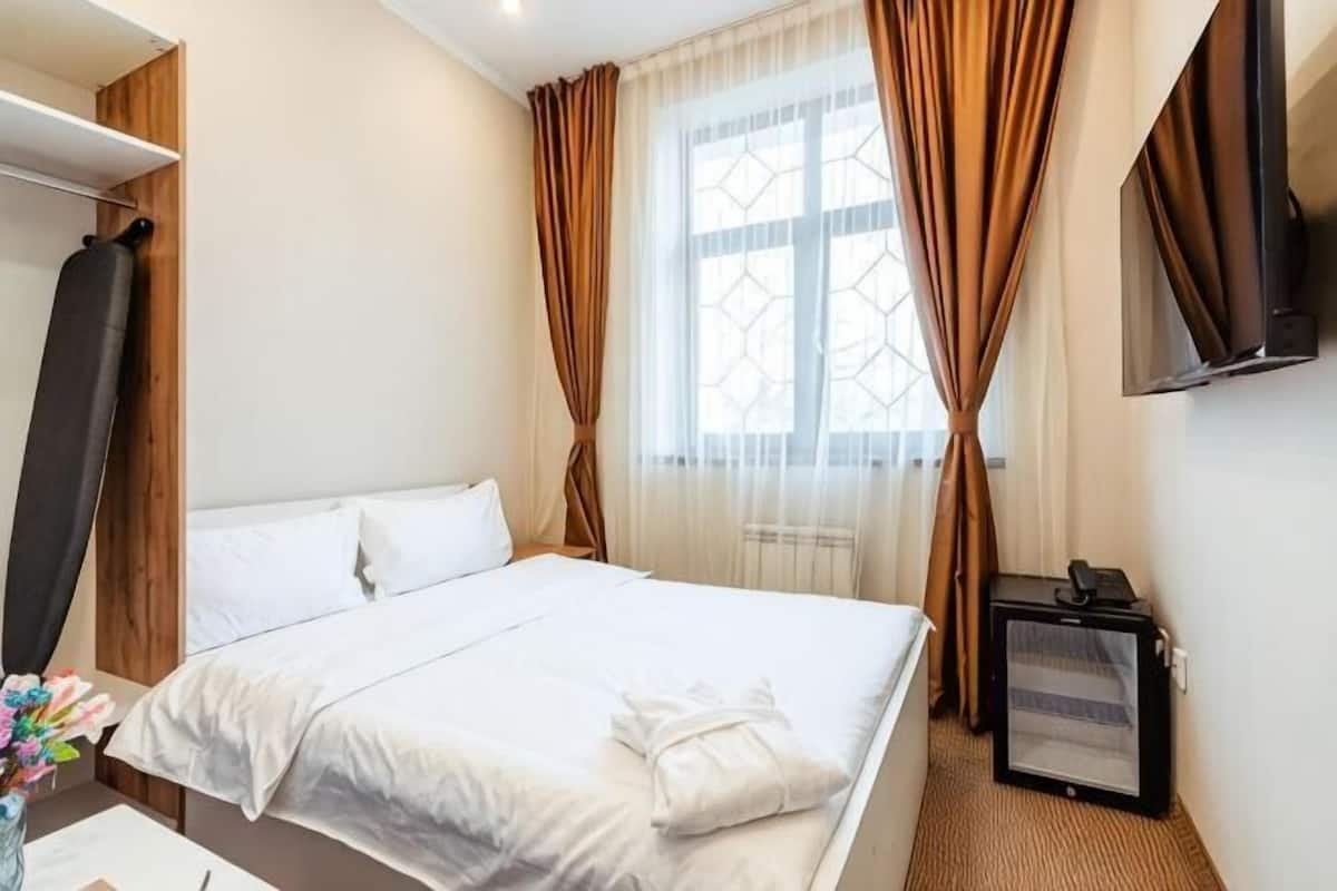 A hotel room with a bed, a TV, a window with curtains, and a desk with a computer.
