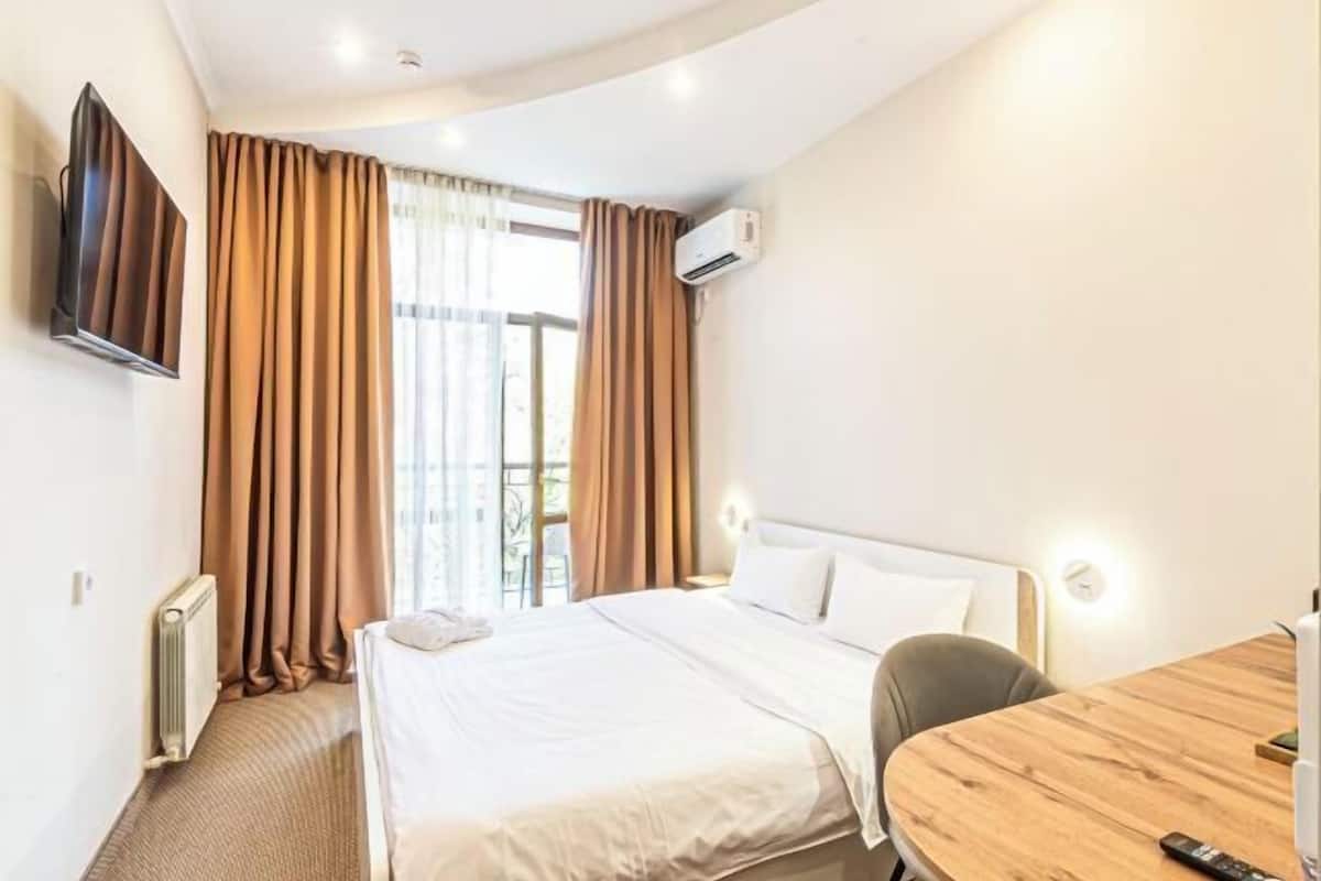 A hotel room with a bed, a wooden table, a TV mounted on the wall, and an air conditioning unit.