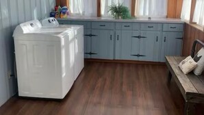 A laundry room with a white washer and dryer, wooden cabinets, and a window with curtains. - Cozy Ranch Retreat Near Jefferson City - Fun Games, Activities, Sports, & Land! (Loose Creek)
