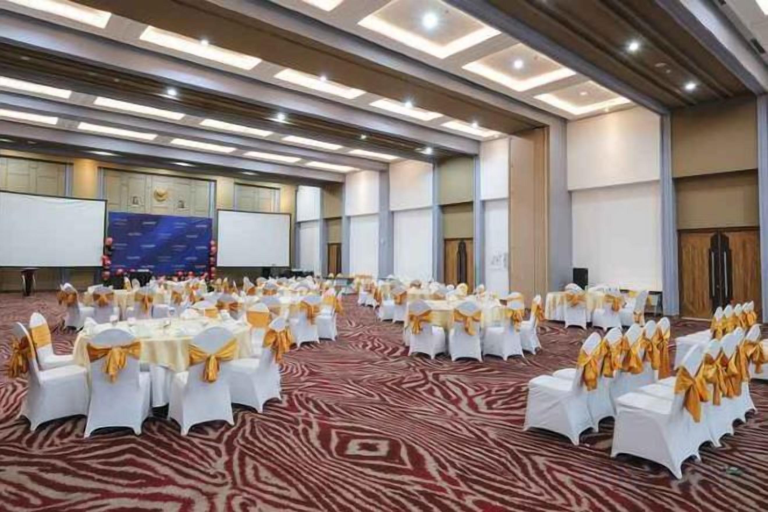 Un amplio salón de conferencias con filas de mesas y sillas blancas, un escenario y una gran pantalla.