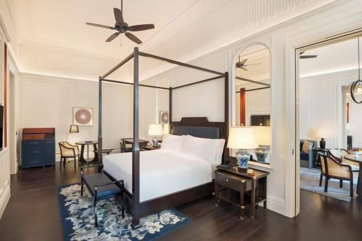 A four-poster bed with a ceiling fan, a desk with a lamp, and a chair in a hotel room with dark wood flooring and white walls.