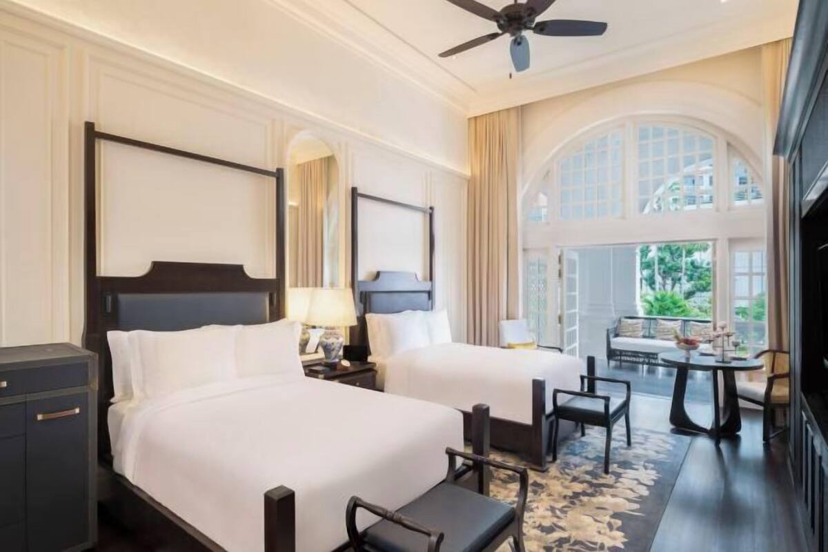 A hotel room with two beds, a desk, a chair, a ceiling fan, and a large window with a view of greenery.