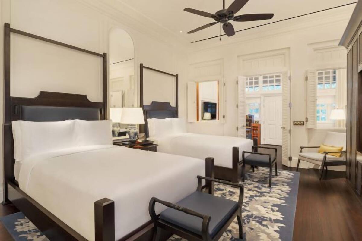 A hotel room with two beds, a ceiling fan, a chair, and a window with shutters.