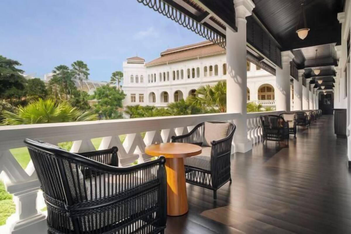 A covered outdoor area with wicker furniture and a round table, overlooking a white building with arches and a garden.