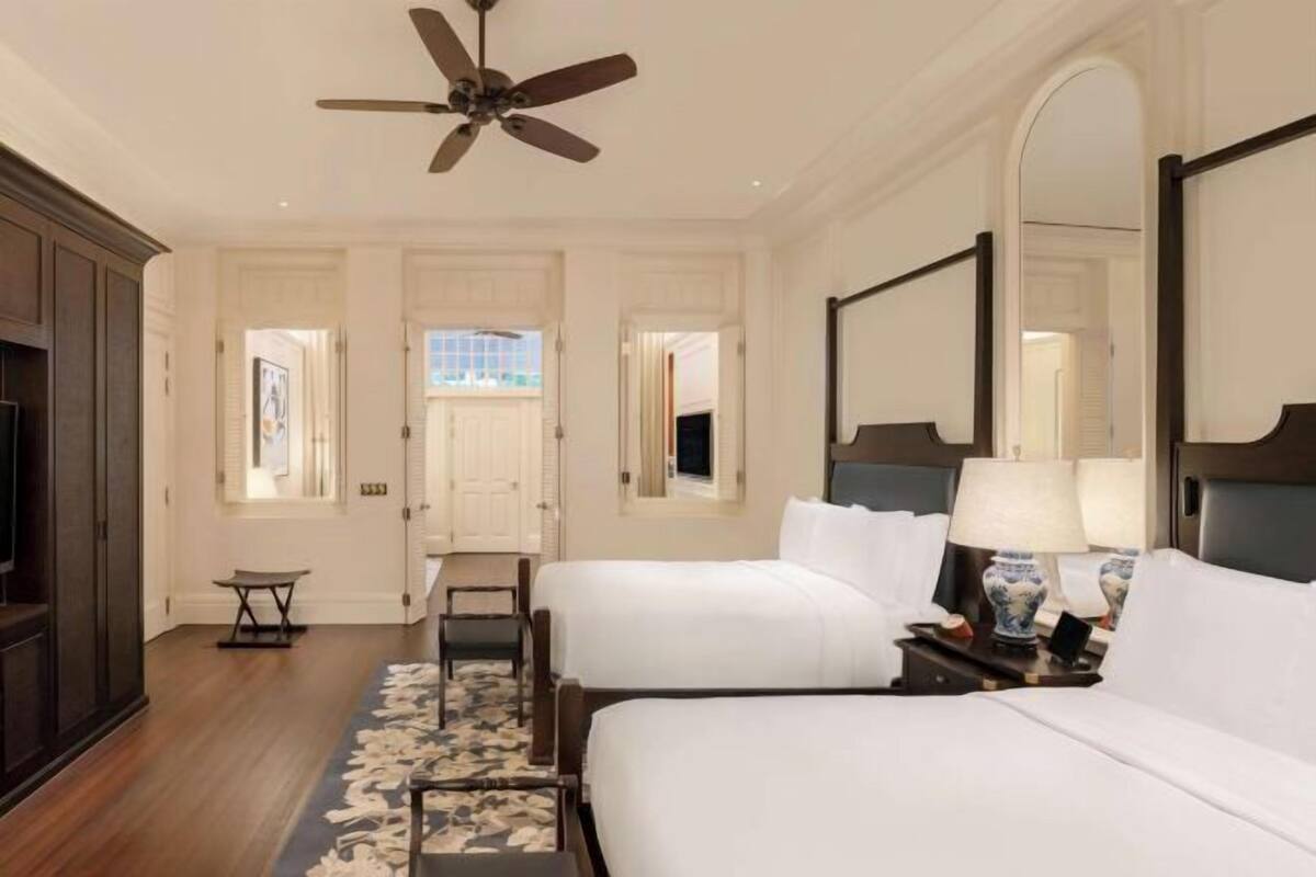 A hotel room with two beds, a ceiling fan, a large mirror, and a view of the outside.