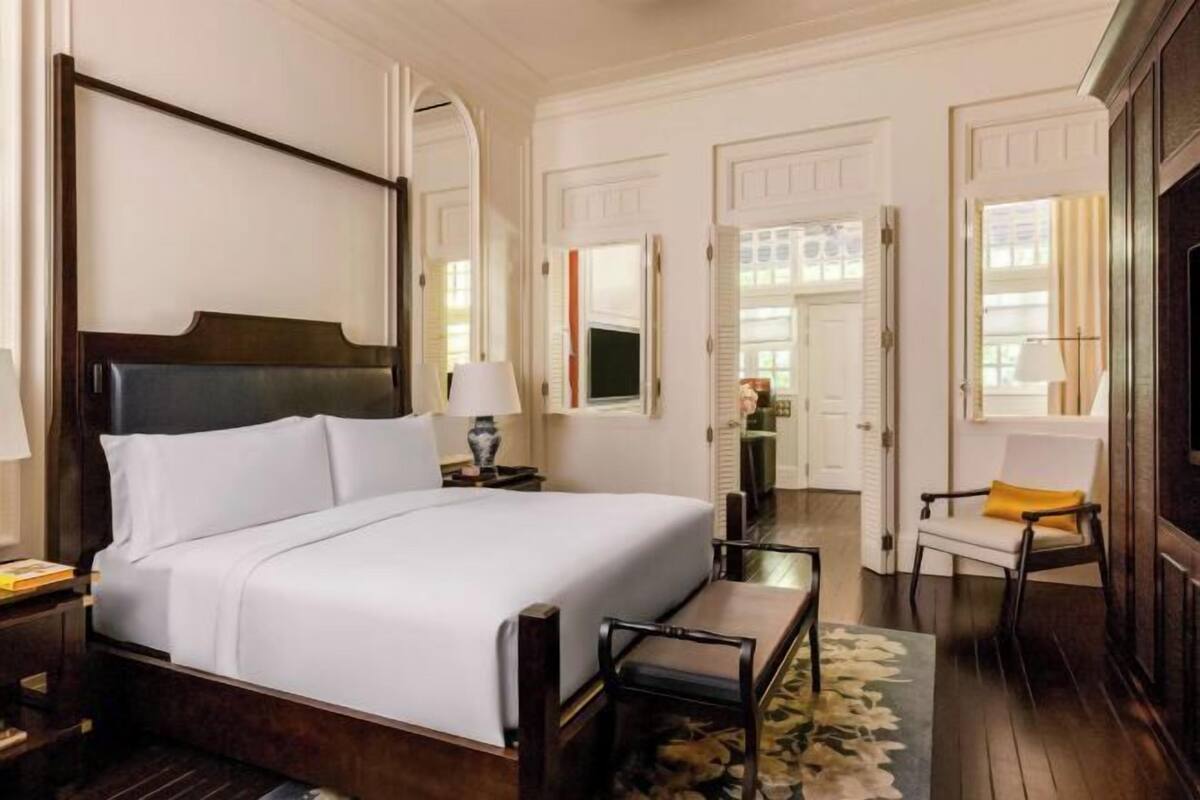 A hotel room with a large bed, a bedside table with a lamp, a chair with a yellow cushion, and a view of the hallway.