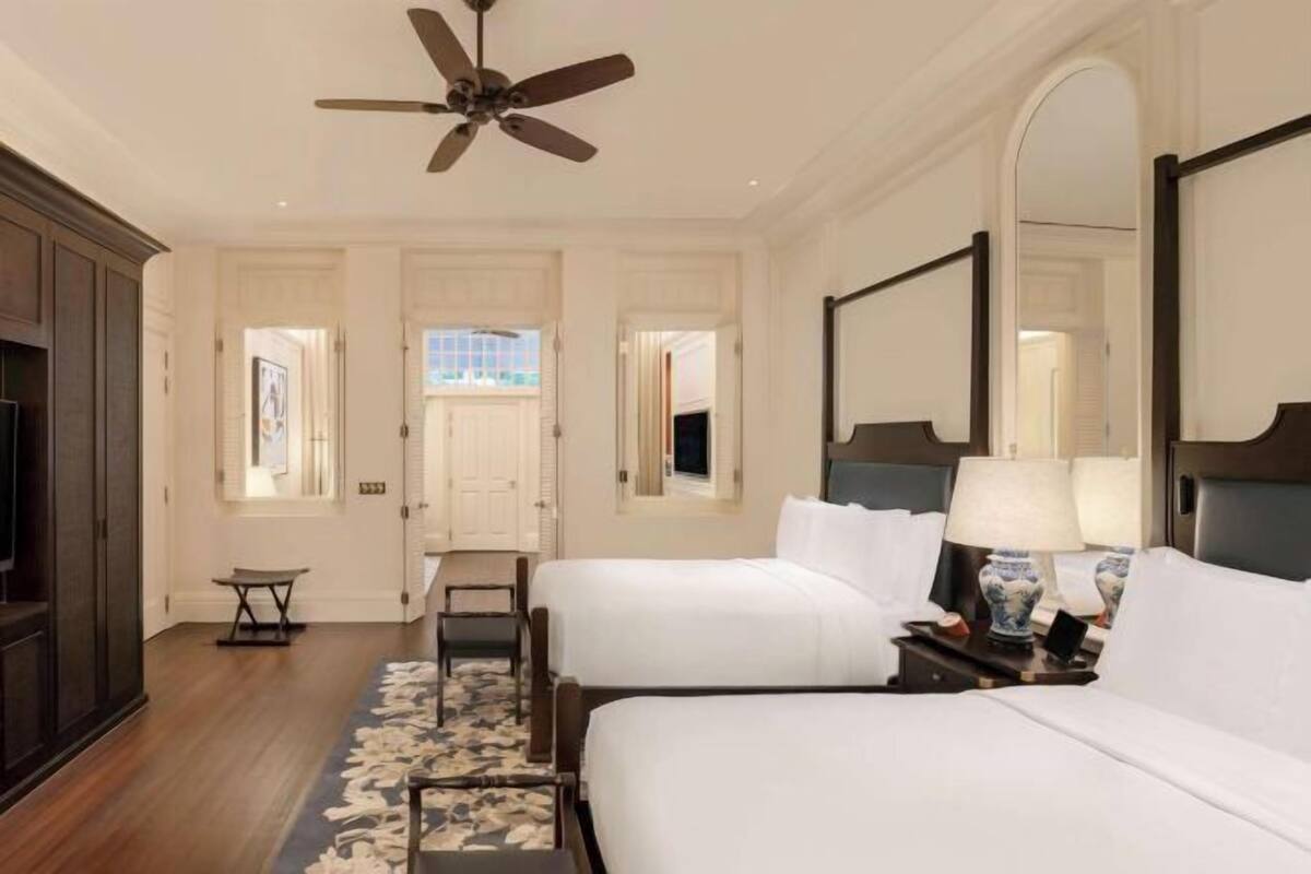 A hotel room with two beds, a ceiling fan, a large mirror, a nightstand with a lamp, and a wooden floor.