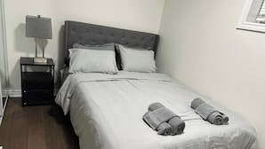 A neatly made bed with a grey headboard and white bedding, a bedside table with a lamp, and two rolled towels on the bed. - Ajax City Suite (Ajax)