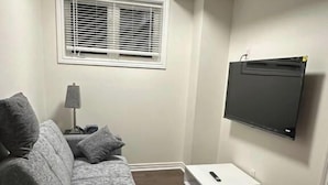 A small, well-lit room with a grey couch, a white side table, a lamp, a mounted flat-screen TV, and a window with blinds. - Ajax City Suite (Ajax)