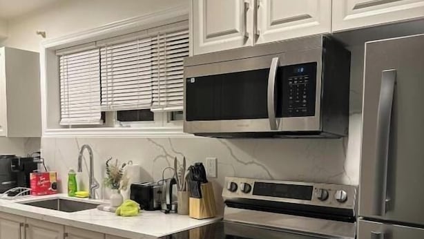 A modern kitchen with white cabinetry, a stainless steel refrigerator, an oven, a microwave, and a sink with a window above. - Ajax City Suite (Ajax)