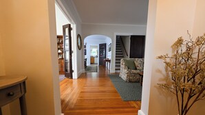 Un pasillo con piso de madera, una escalera y un área de sala con un sofá y un jarrón de flores secas.