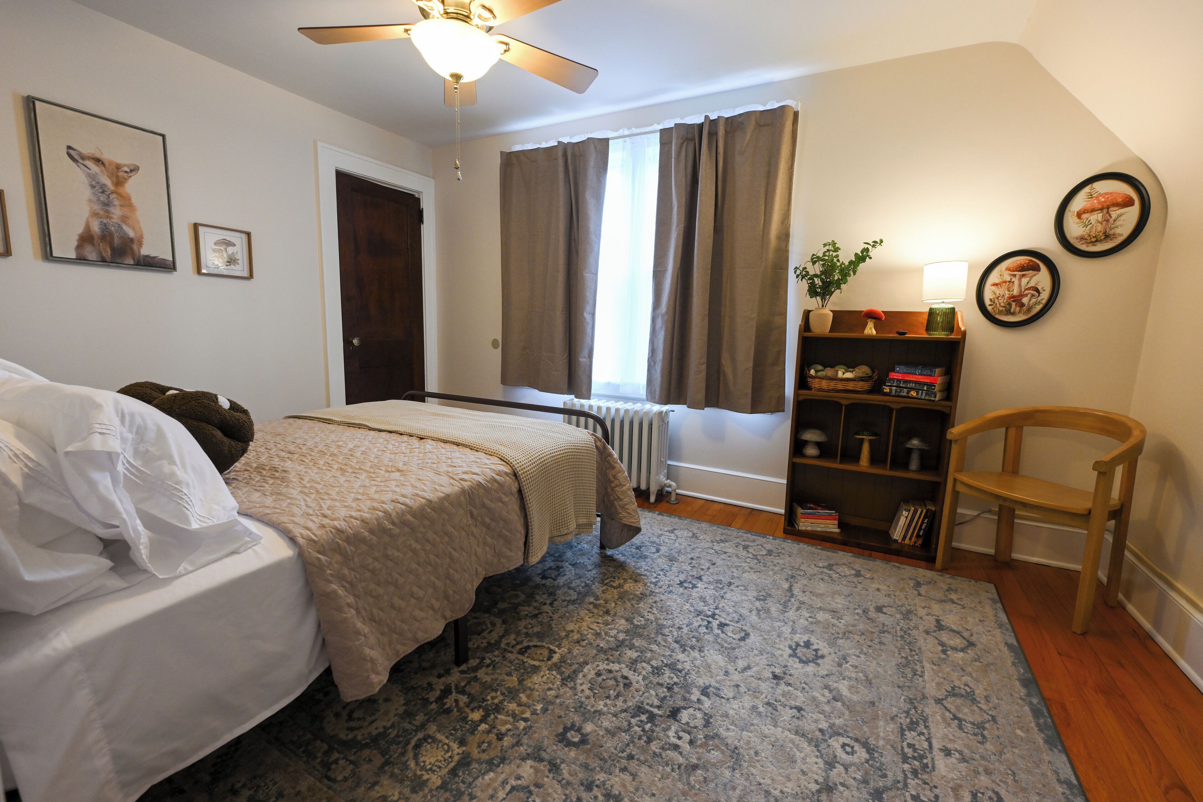 A bedroom with a bed, a radiator, a bookshelf, a chair, and a window with curtains.