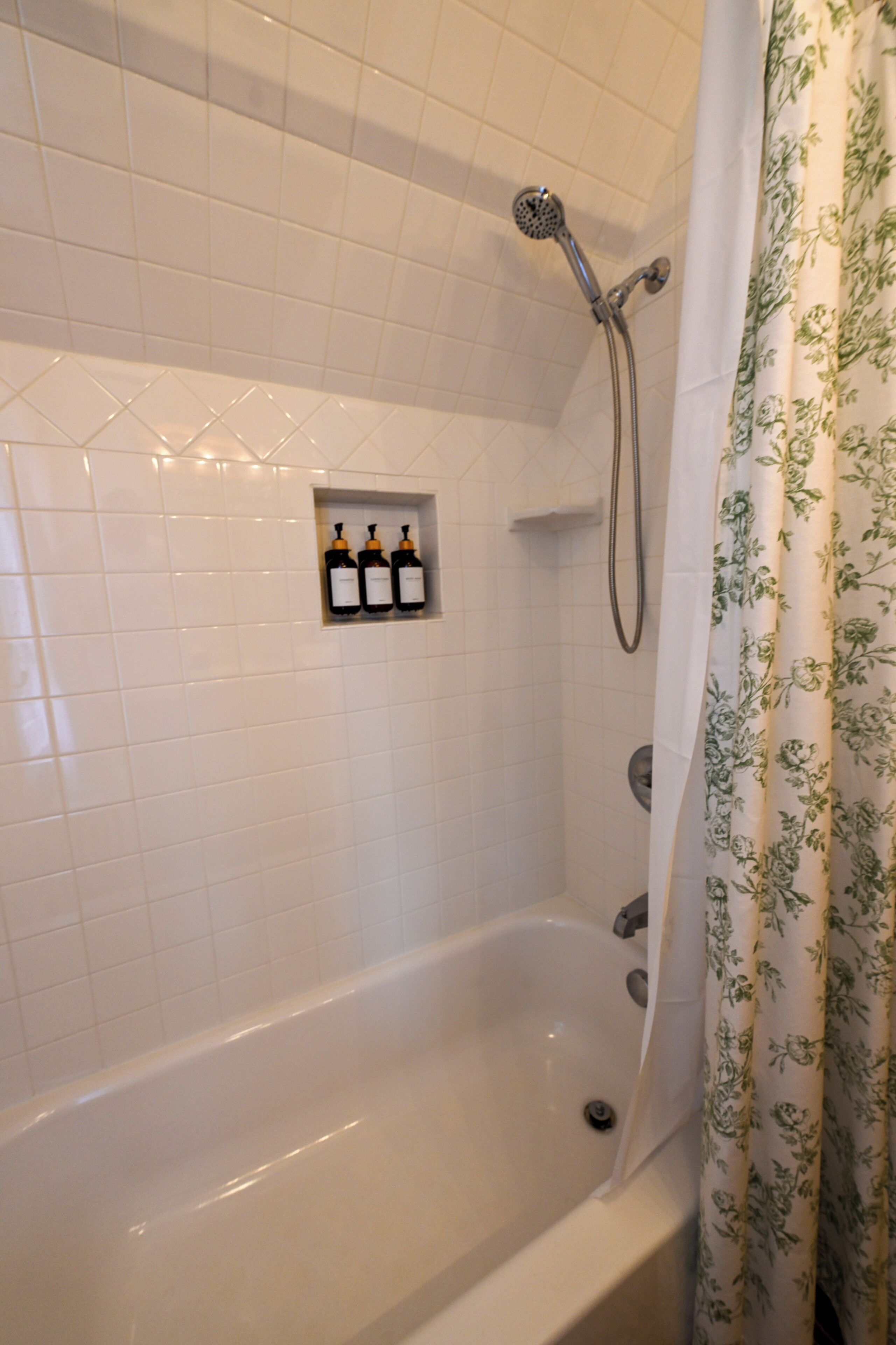 A bathroom with a bathtub, a showerhead, and a floral shower curtain. There are three bottles of shampoo or conditioner on the wall.