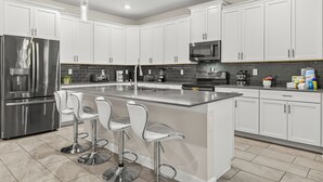 A modern kitchen with white cabinets, stainless steel appliances, and a central island with bar stools. - 6BR Disney Retreat w/ Pool, Game Loft & Resort Amenities (Kissimmee)