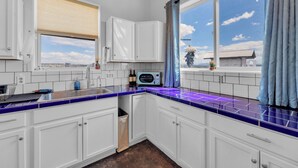 A modern kitchen with white cabinets, a blue and purple tiled backsplash, a window with a view of buildings, and a microwave on the counter. - Starry Sky + Serene Setting: Rural New Mexico Gem (Carrizozo)