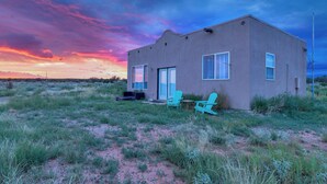 A single-story building with a flat roof, large windows, and a door. There is a small outdoor seating area with two chairs and a table. - Starry Sky + Serene Setting: Rural New Mexico Gem (Carrizozo)