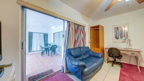 Une pièce avec un canapé en cuir bleu, un bureau en bois avec une chaise, une porte-fenêtre coulissante donnant sur une terrasse extérieure avec une table et des chaises, et un ventilateur de plafond.