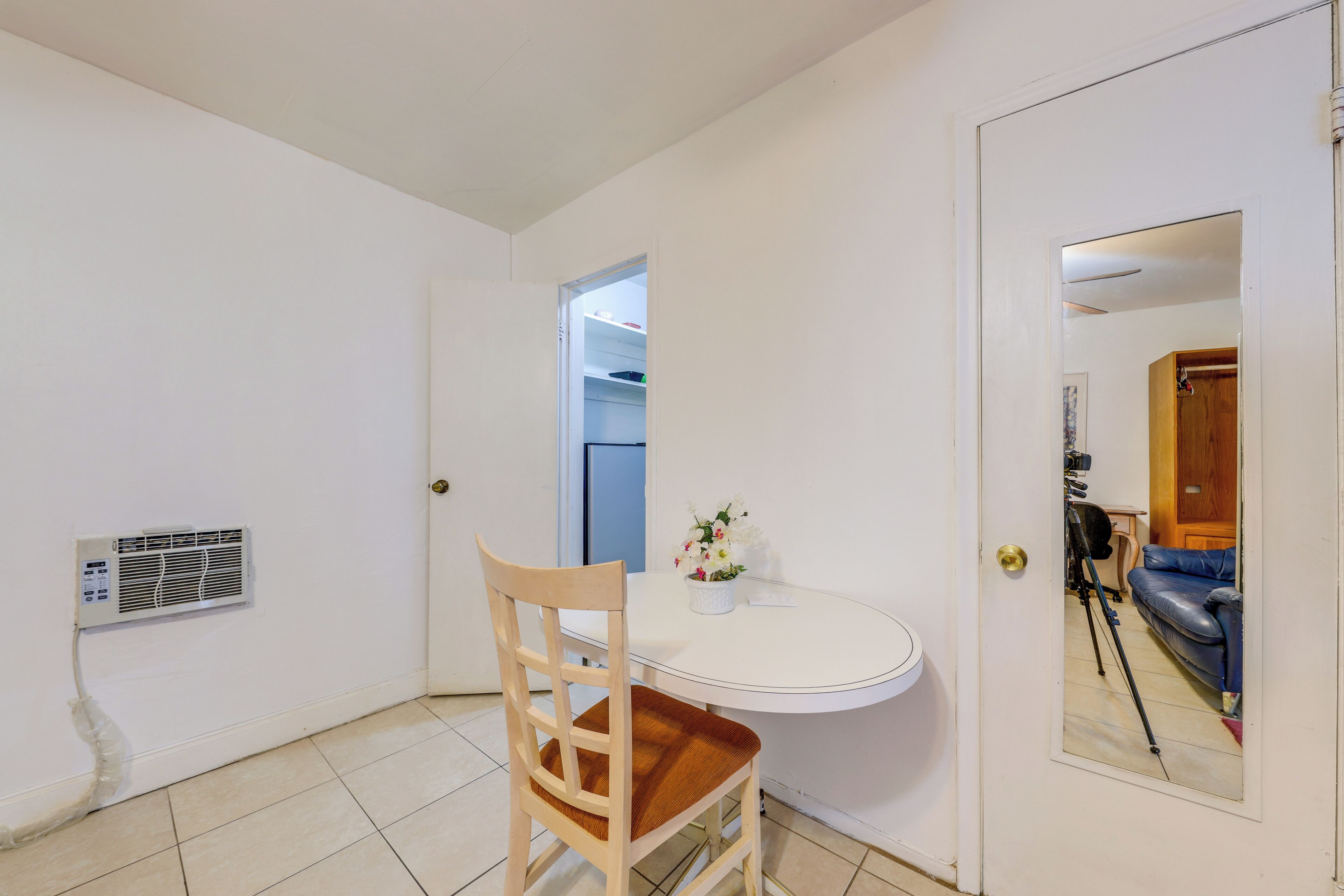 Quarto pequeno com uma mesa branca, uma cadeira de madeira, um aparelho de ar condicionado branco na parede e uma porta com um espelho refletindo um sofá azul e um tripé de câmera.