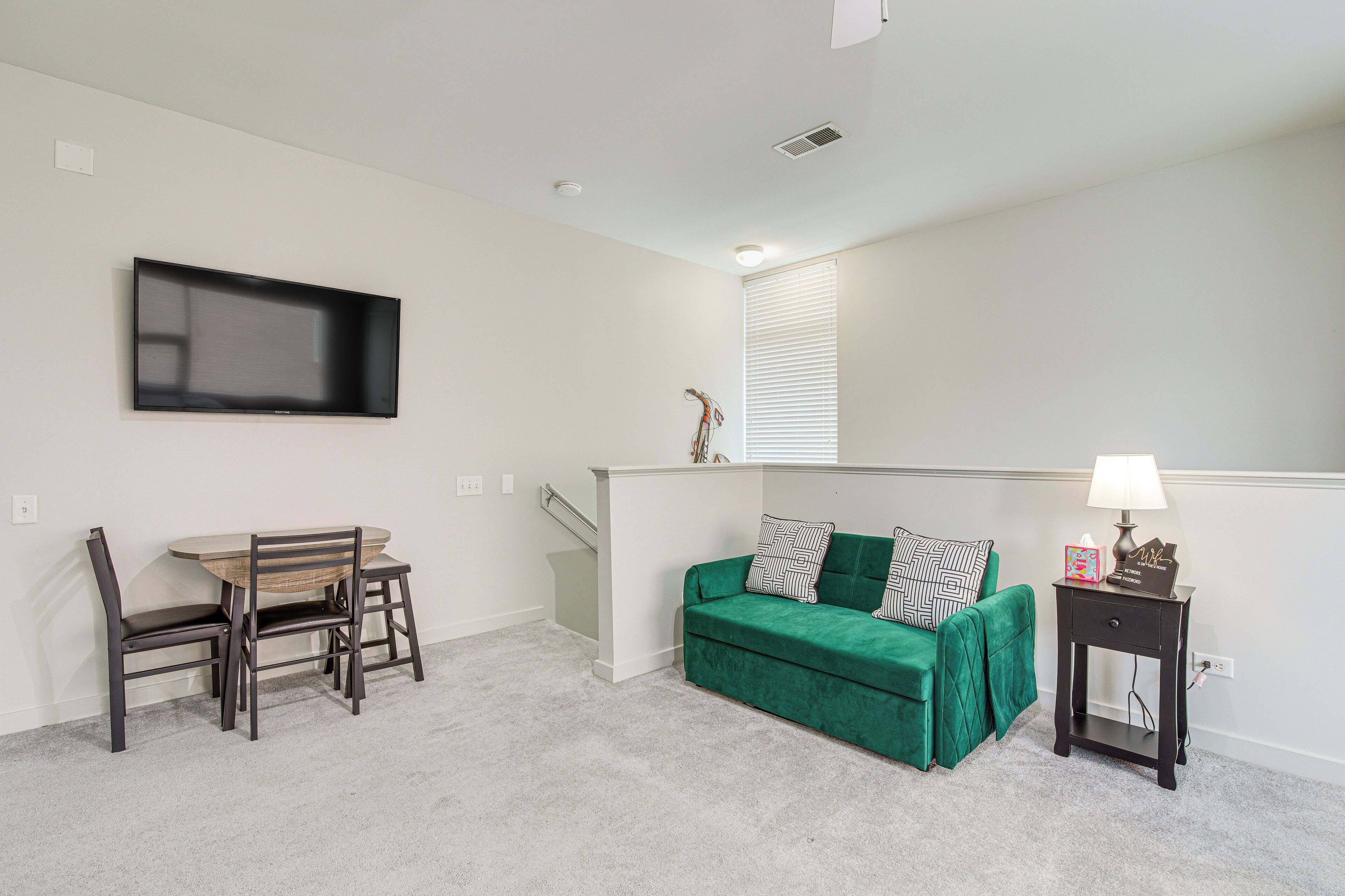 A living room with a green sofa, a wooden dining table with chairs, a TV mounted on the wall, and a small side table with a lamp.