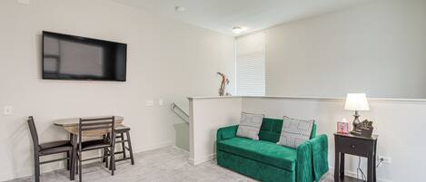 A living room with a green sofa, a wooden dining table with chairs, a TV mounted on the wall, and a small side table with a lamp.