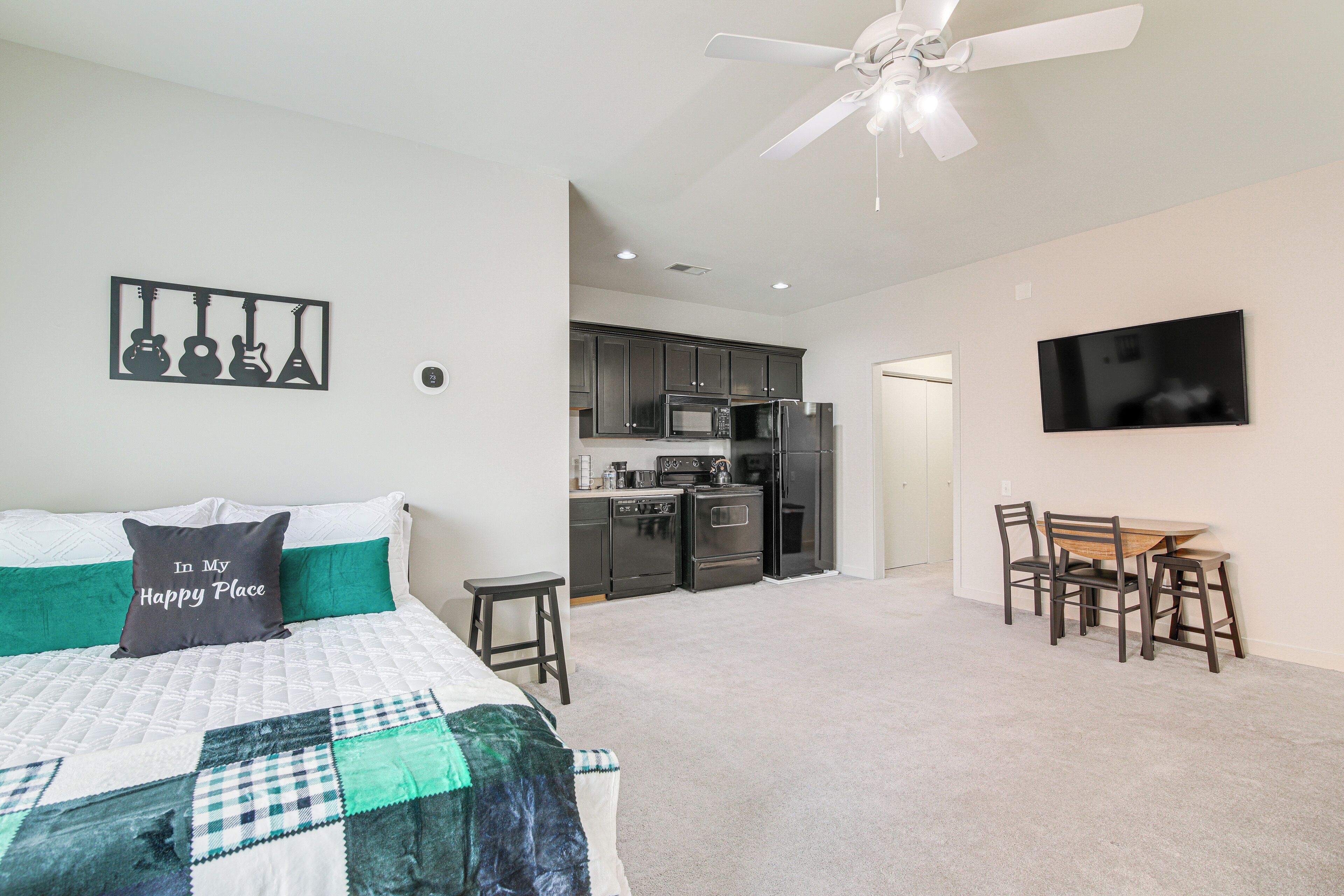 A bedroom with a bed, a quilt, a pillow, a wall-mounted guitar decoration, a ceiling fan, and a kitchen area with a dining table and chairs.