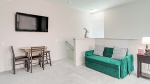 A room with a green sofa, a dining table with chairs, a flat-screen TV mounted on the wall, and a staircase leading to an upper level.