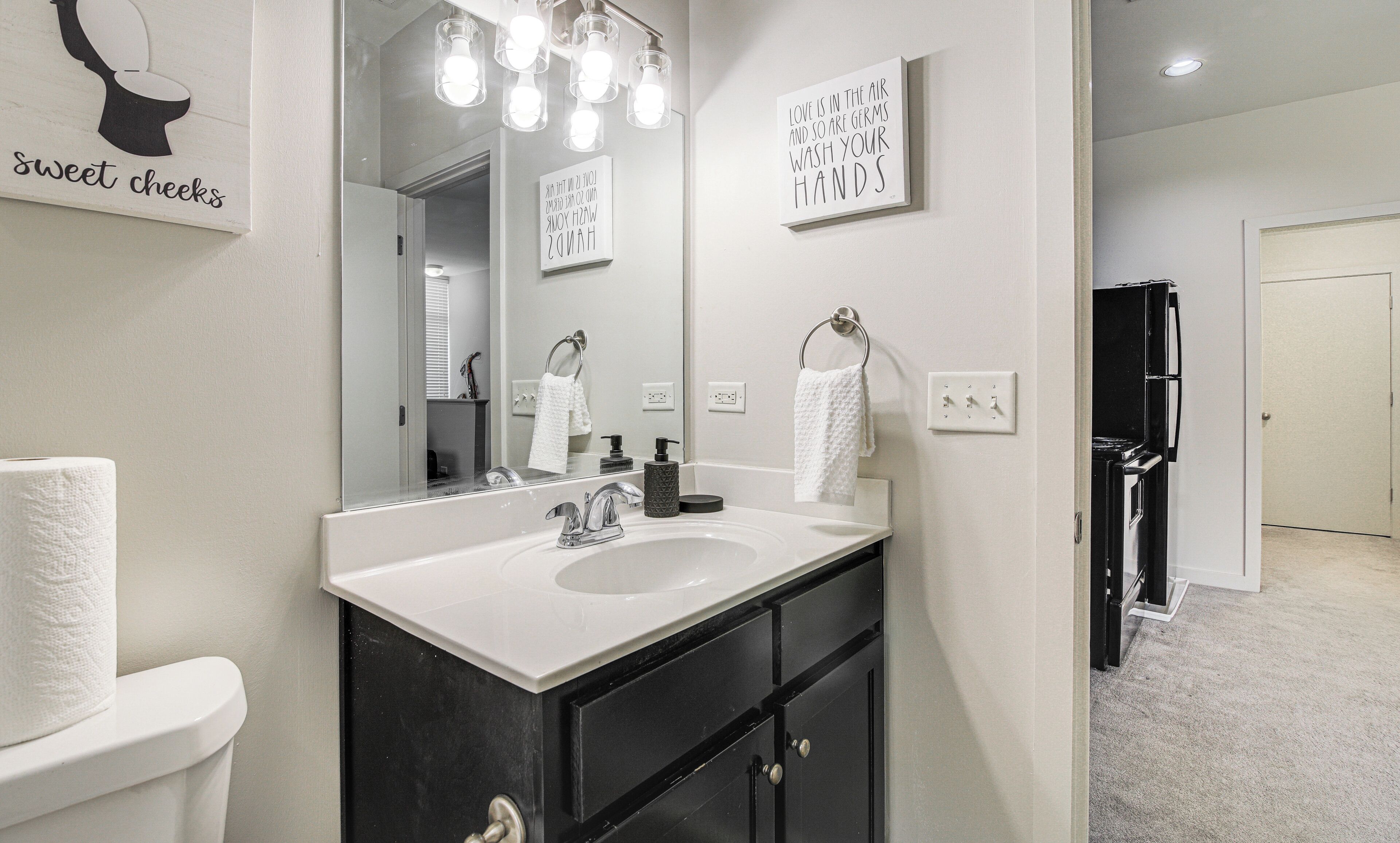 A modern bathroom with a white sink, black cabinets, and a large mirror. There are wall-mounted lights, a towel rack, and a sign that reads "Love is in the air and so are germs wash your hands."