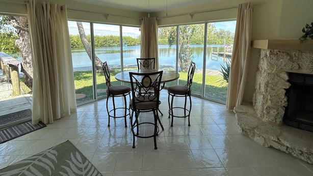 Un vaste salon doté d’une cheminée en pierre, un coin repas avec une table et des chaises, de grandes fenêtres offrant une vue sur un lac et un ventilateur de plafond.