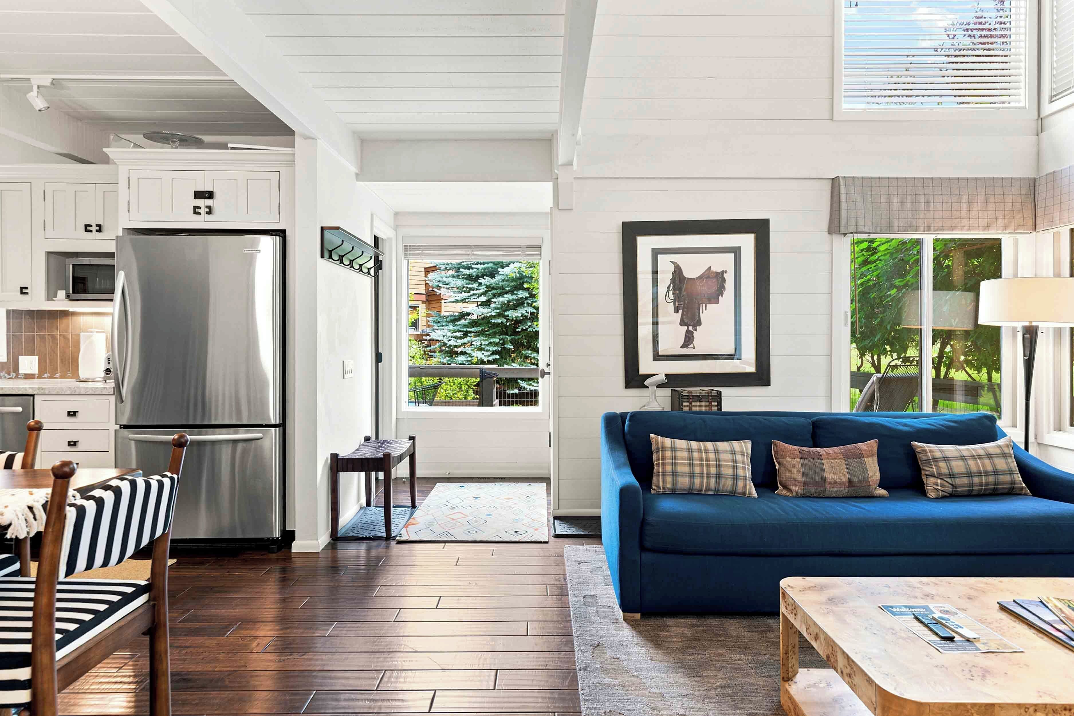 A modern kitchen with a stainless steel refrigerator, a dining area with striped chairs, a blue sofa with plaid cushions, and a view of greenery outside.