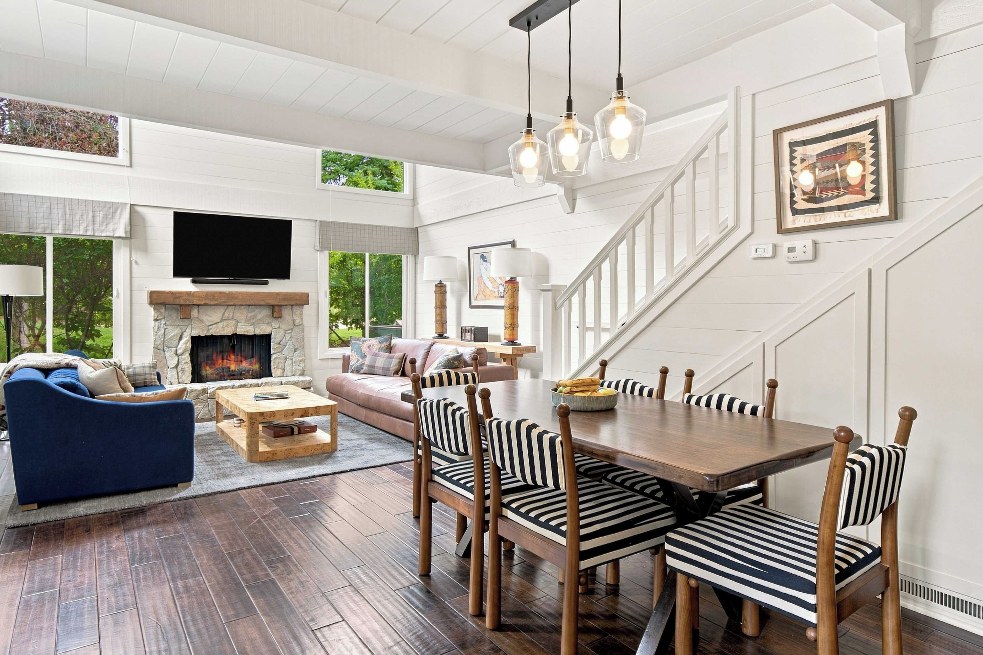 A spacious living area with a fireplace, a television mounted above it, and a dining area with striped chairs and a wooden table.