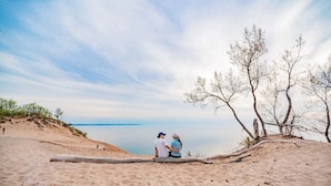 A sandy beach with two people sitting on a log, overlooking a calm body of water.