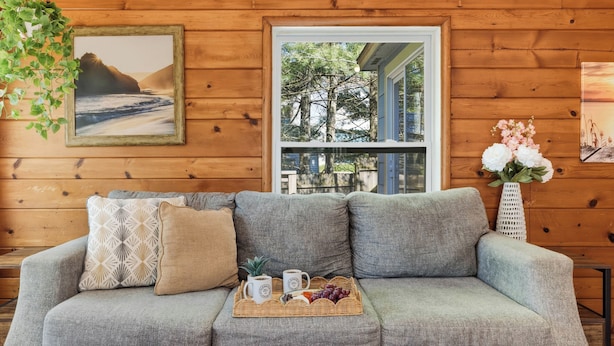 A grey sofa with patterned cushions, a tray with coffee mugs and fruit, and a framed picture of a coastal landscape.