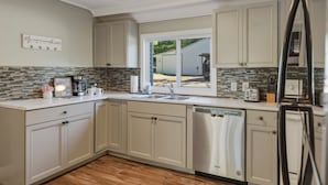 A kitchen with light-colored cabinets, a dishwasher, a refrigerator, and a window with a view of a backyard.