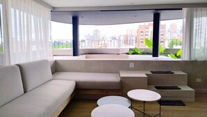 A modern living room with a sofa, two round tables, and a view of a cityscape through large windows. - Node Hotel (Manizales)