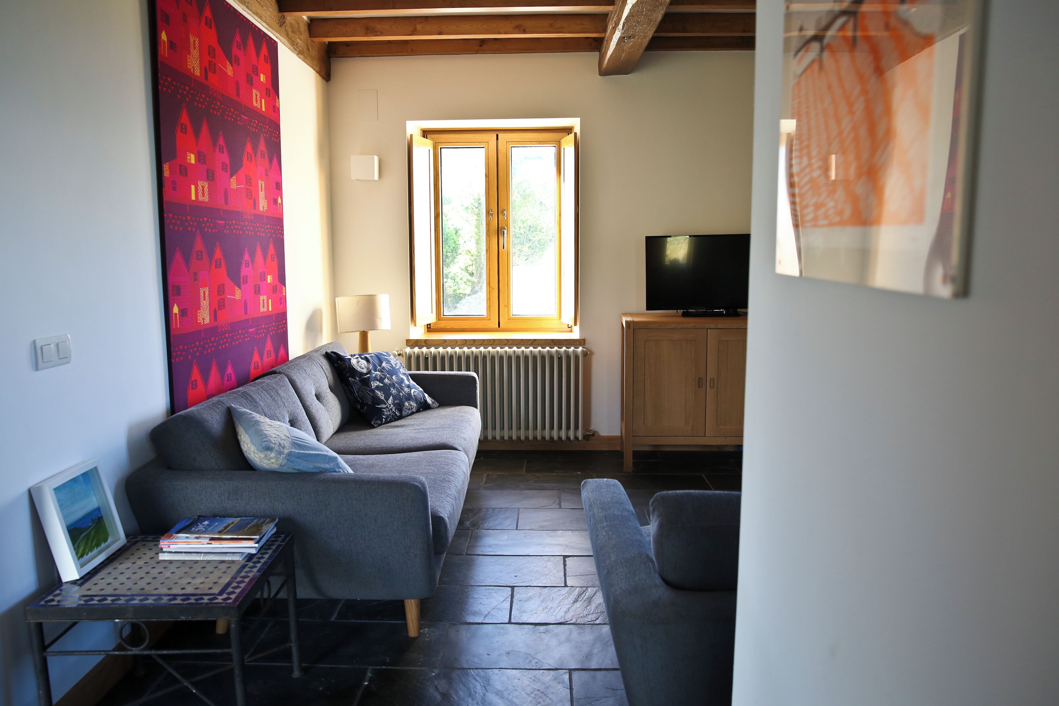 A living room with a grey sofa, a small table with books, a radiator, a wooden cabinet, and a television.