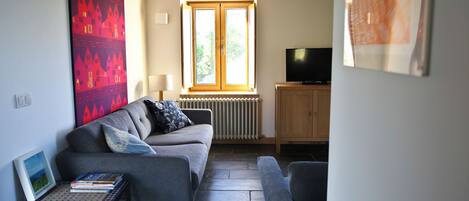 A living room with a grey sofa, a small table with books, a radiator, a wooden cabinet, and a television.