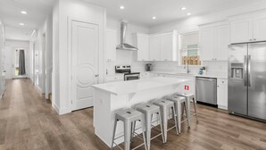 A modern kitchen with white cabinetry, a central island with bar stools, stainless steel appliances, and a wooden floor. - Ellis Sands with LSV, Arcade, and Billiards (Miramar Beach)