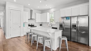 A modern kitchen with white cabinetry, stainless steel appliances, and a central island with bar stools. - Ellis Tide with LSV, Arcade, and Billiards (Miramar Beach)