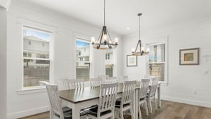 Une salle à manger avec une grande table en bois et des chaises blanches, deux lustres et de grandes fenêtres équipées de stores.