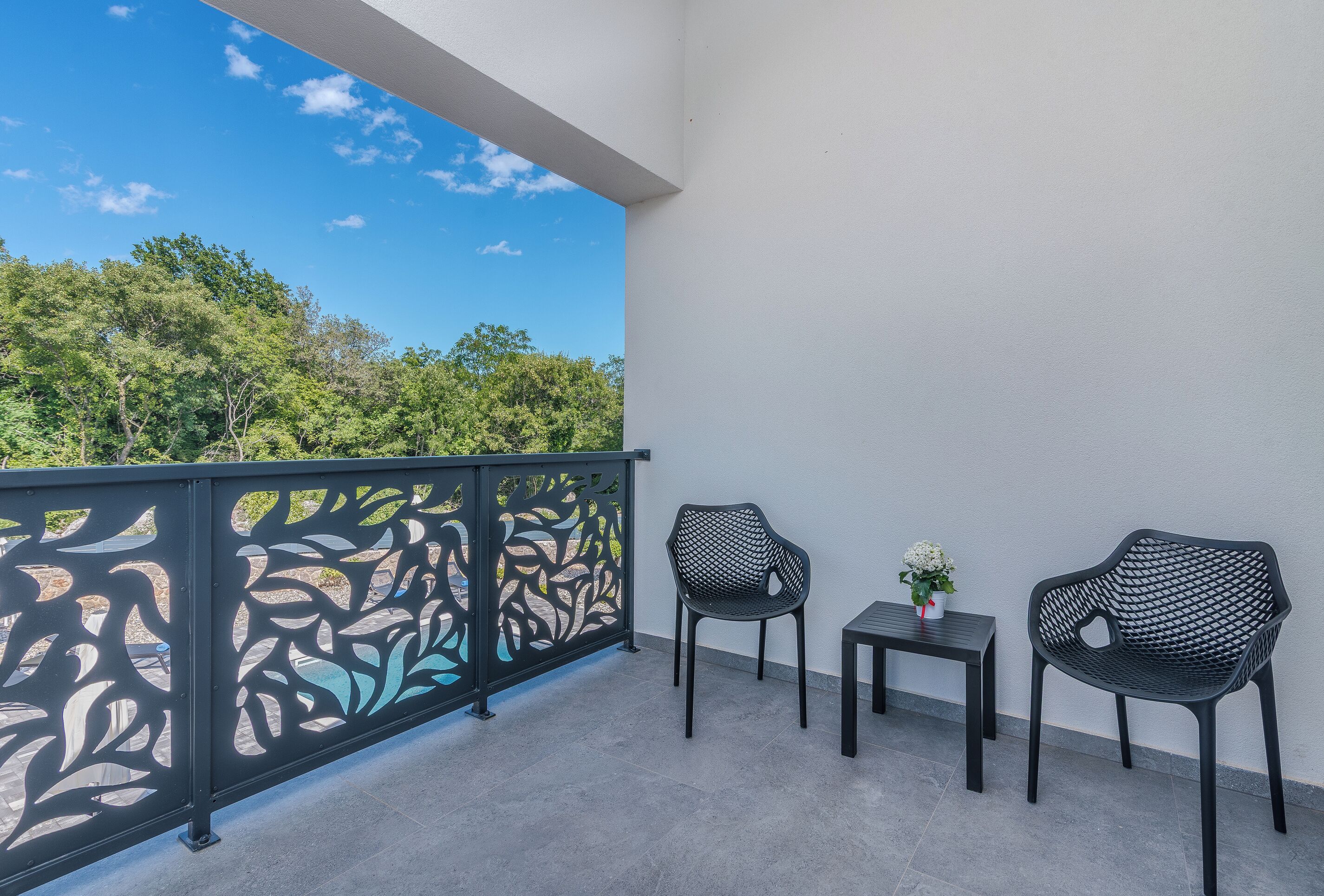 A balcony with two chairs and a small table, a decorative metal fence, and a view of trees and sky.
