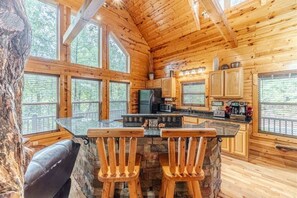 A cabin interior with a stone fireplace, wooden bar stools, a kitchen area, and large windows.