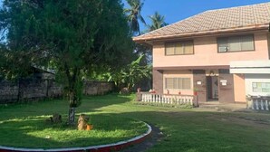 A two-story building with a tiled roof, a front entrance, and a small garden area with a tree and a lawn. - WJV INN LILOAN (Liloan)
