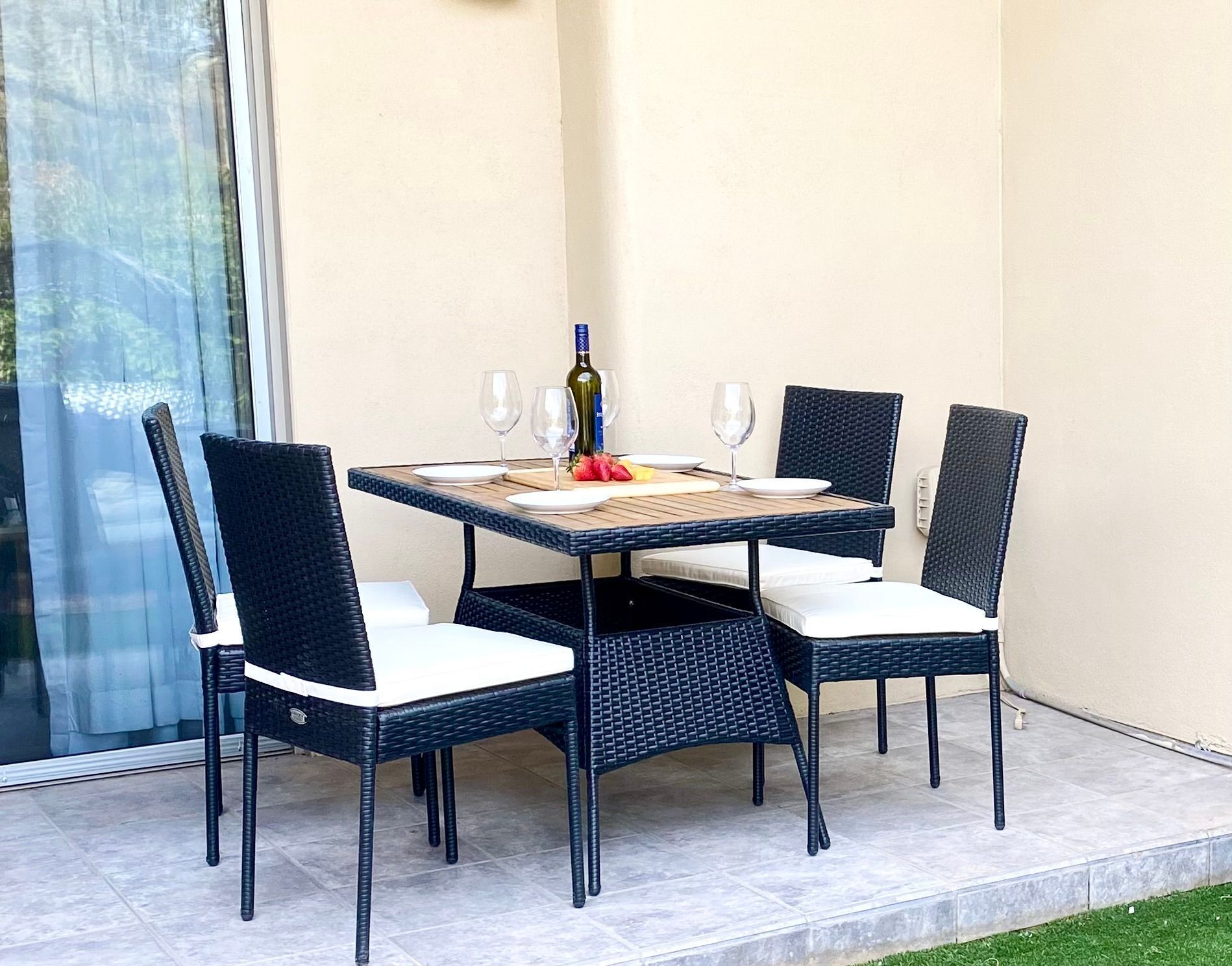 Una mesa de comedor con cuatro sillas, una botella de vino y dos copas en un balcón con puerta de cristal y suelo de baldosas.