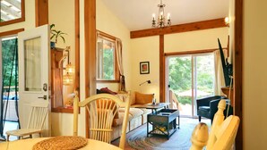 A cozy living room with a wooden floor, a sofa, a dining table with woven placemats, and a chandelier. - Welcome to the Meadowverse, your peaceful retreat (Galiano Island)