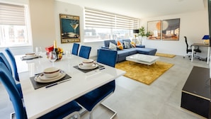 A modern living room with a dining area, featuring a sofa, coffee table, and a dining table set with plates and glasses. - Apartment - Sleeps 6 - Parking - Wifi (Guildford)