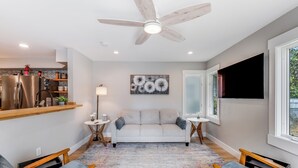 Una sala moderna con un sofá, dos sillones, un ventilador de techo, un televisor y una cocina con un refrigerador y estanterías.