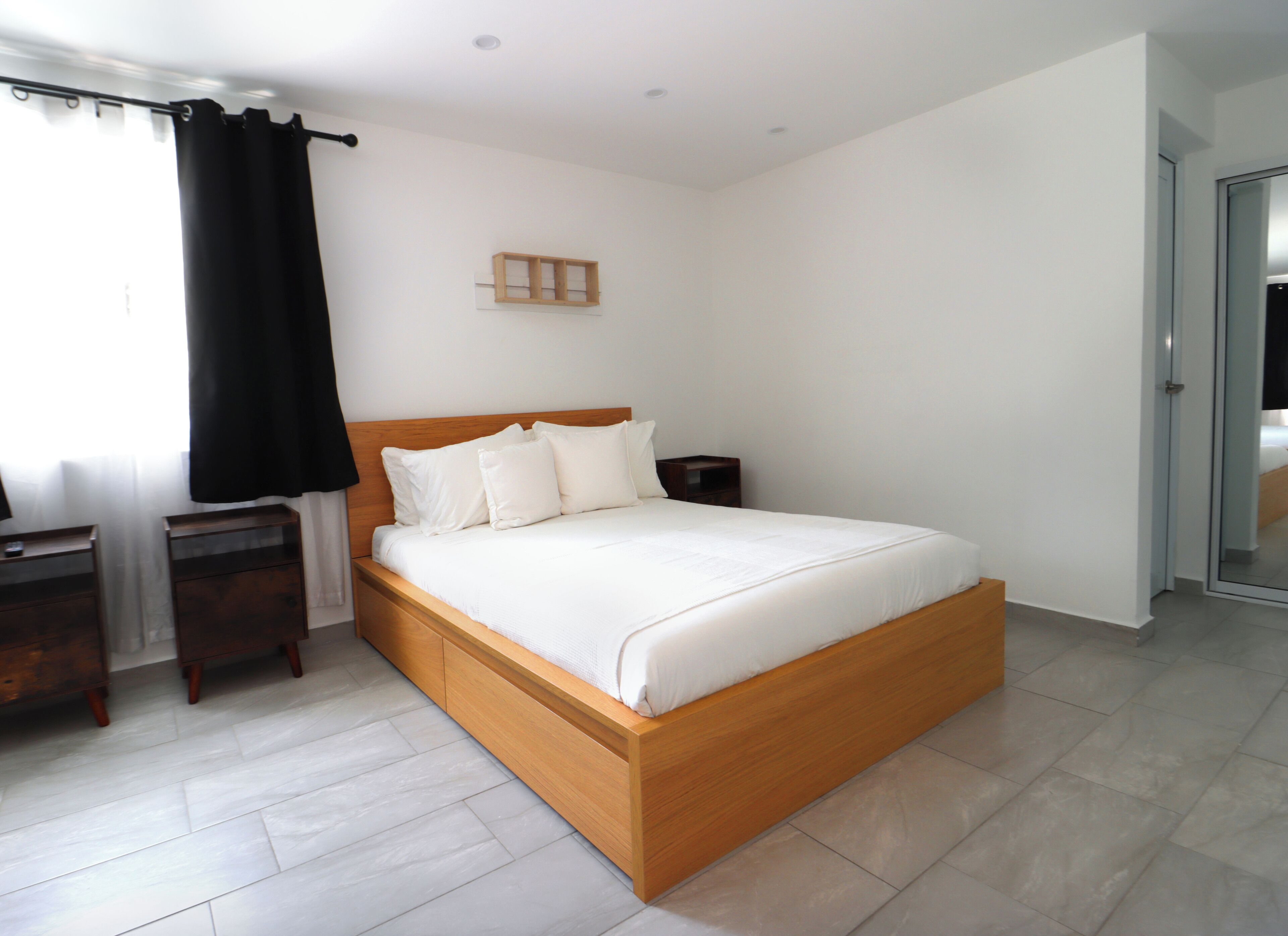 A bedroom with a wooden bed, white bedding, a nightstand, a small wooden shelf, and a window with black curtains.