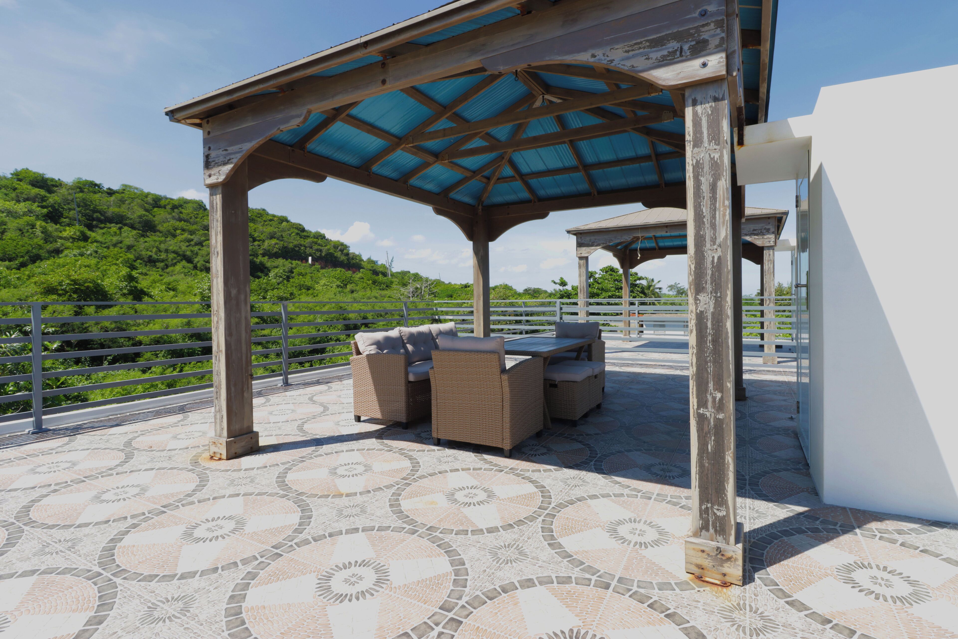 An outdoor patio with a covered seating area, a patterned floor, and a view of a lush green hillside.