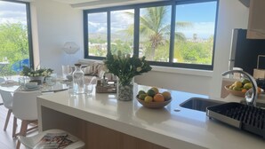 Une cuisine moderne avec une grande fenêtre, un coin repas doté d’une table en verre et un saladier rempli de fruits posé sur le comptoir.