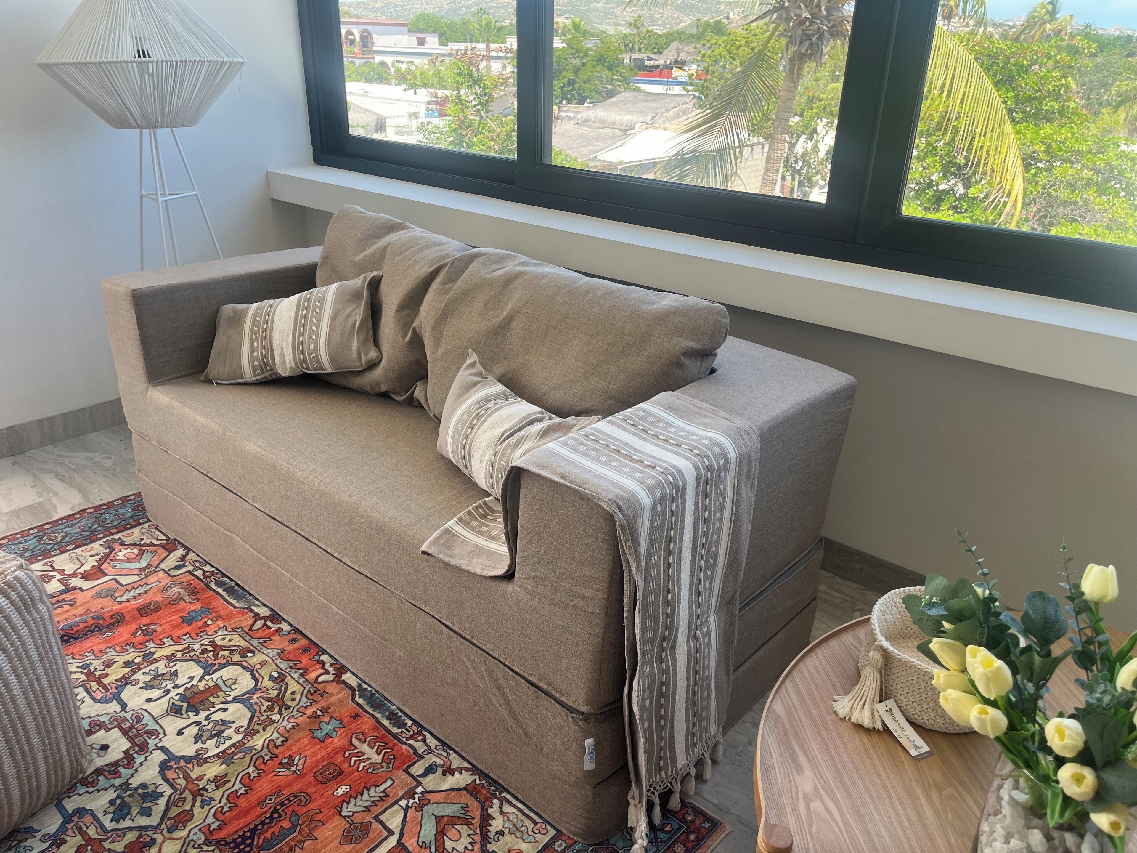 Un salon chaleureux avec un canapé, un tapis à motifs, une petite table ronde ornée d’un vase de fleurs et une vue sur un quartier résidentiel par la fenêtre.